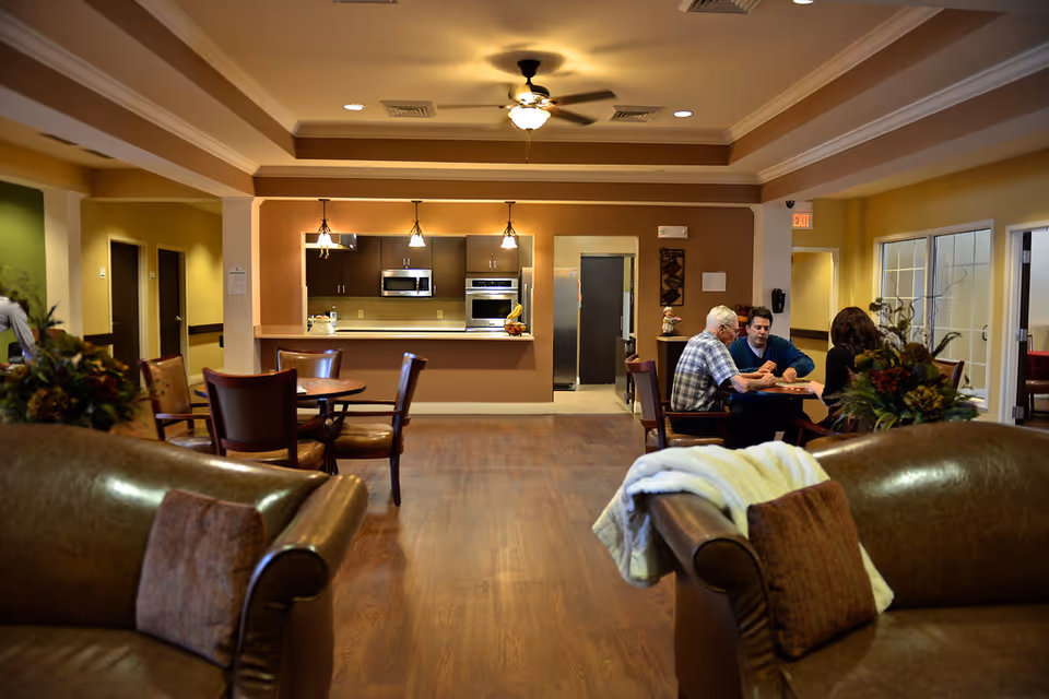 A cozy senior living common area with brown leather sofas and pillows in the foreground, a wooden floor, and a kitchen area with hanging lights in the background. Three people are seated at a table engaged in conversation near the kitchen.