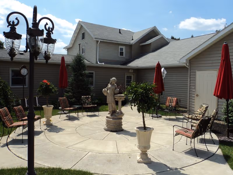 Sunlit circular courtyard with a central statue, potted plants, chairs and red umbrellas in front of a two-story building.