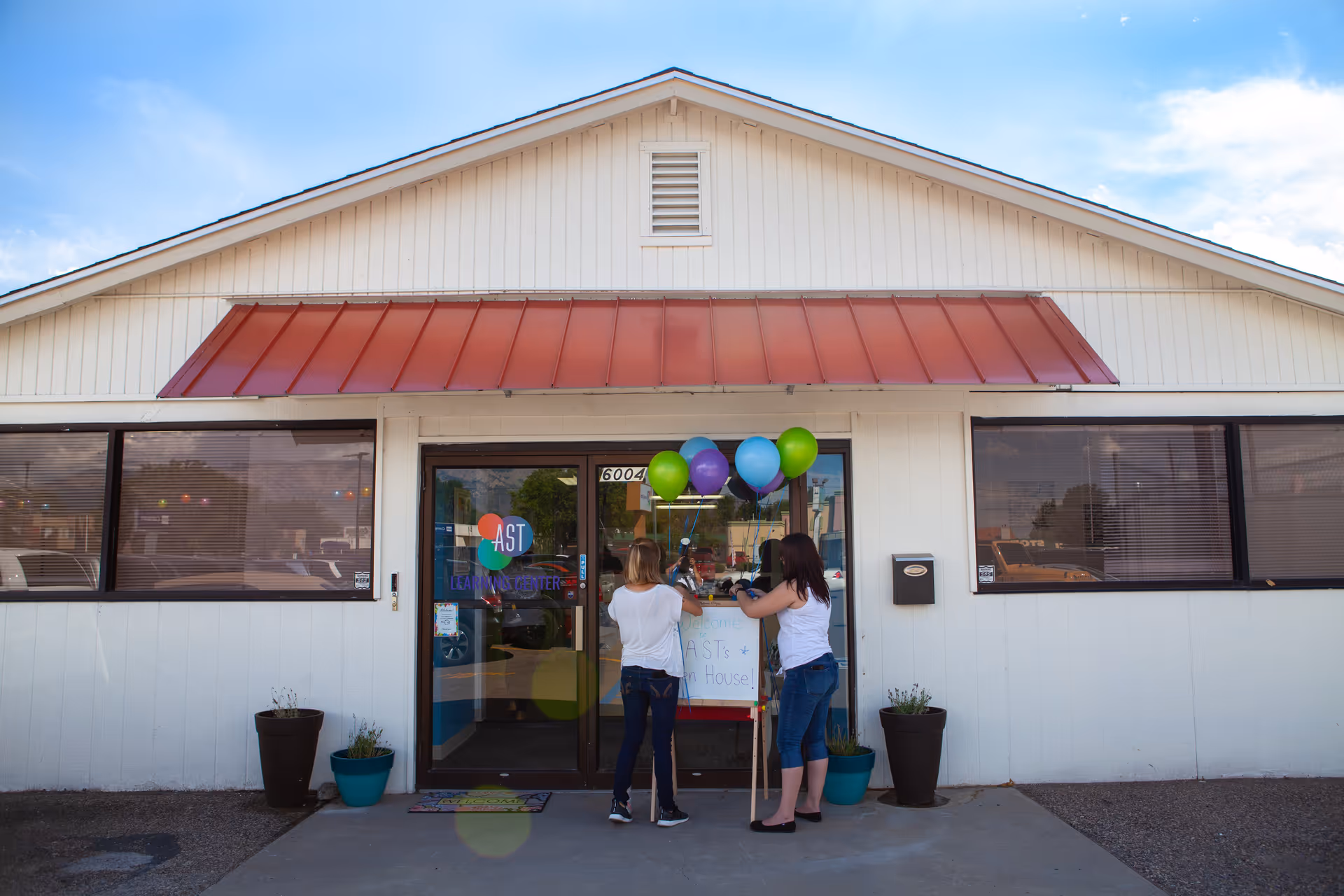 White, single-story building with red awning, balloons at entrance, and two people by the doors.