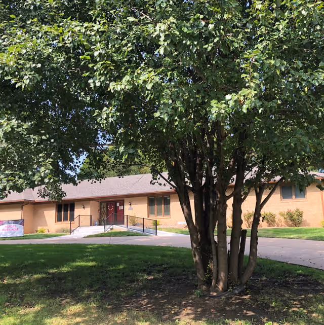 A single-story brick building partially obscured by a large leafy tree in the foreground. The building has a sloped roof and a red double-door entrance with a ramp and handrails. There is a grassy lawn and a paved driveway in front of the building.