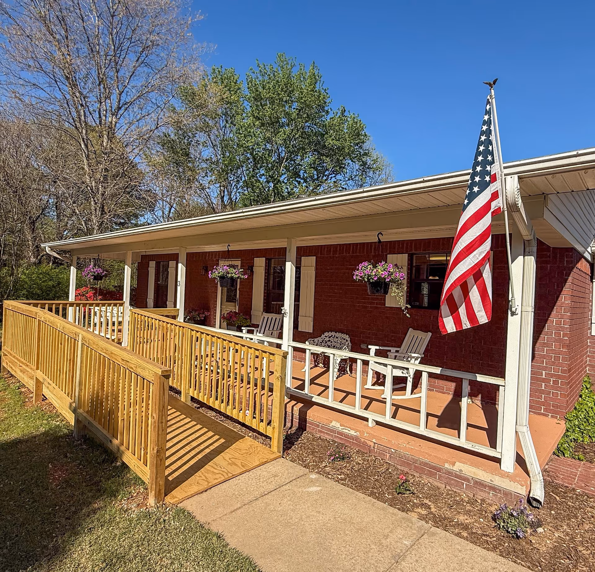 Brick care home exterior with a wooden ramp, porch seating, hanging planters, and American flag.