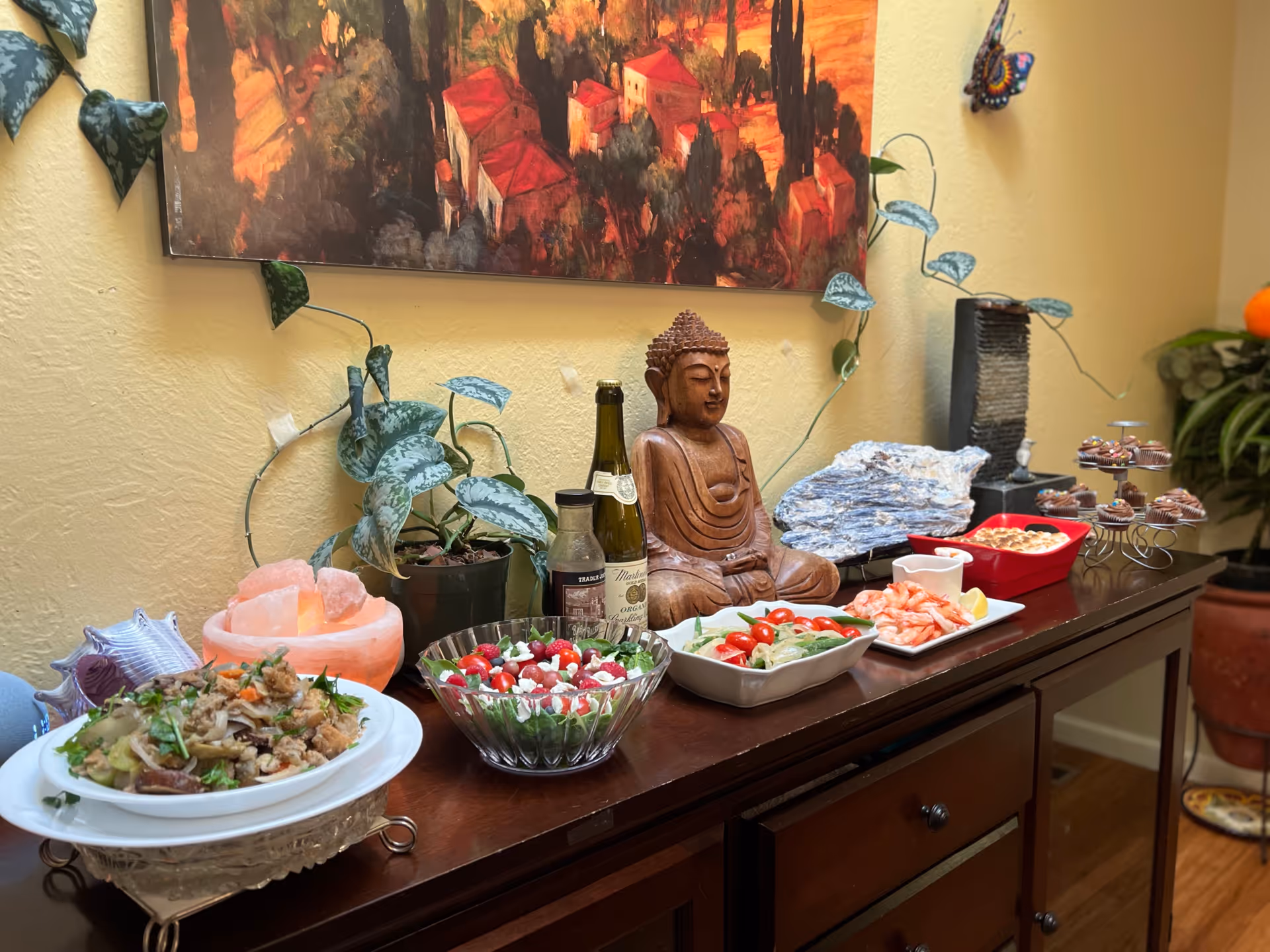 A wooden sideboard displays Buddha statue, salads, shrimp, and desserts amid plants in a dining area.