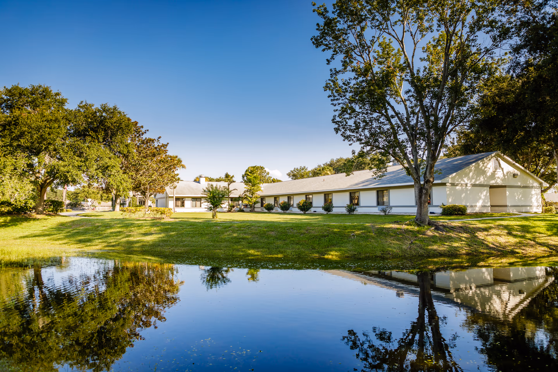 Single-story white senior living facility with a green lawn, trees, and a reflecting pond.
