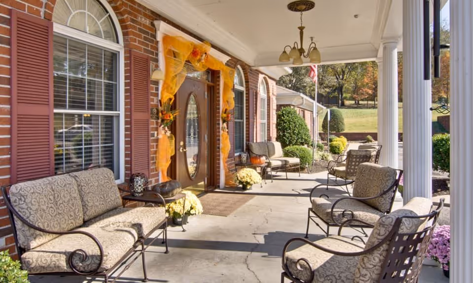 Covered front porch with cushioned metal seating, white columns, and a decorated entry door on a brick building.