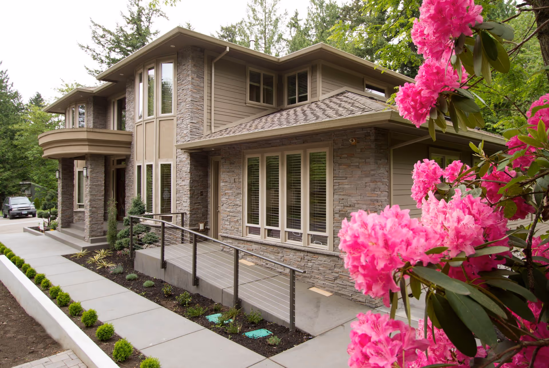 Front exterior of a modern two-story stone-and-siding house with a wheelchair ramp and bright pink flowers in the foreground.