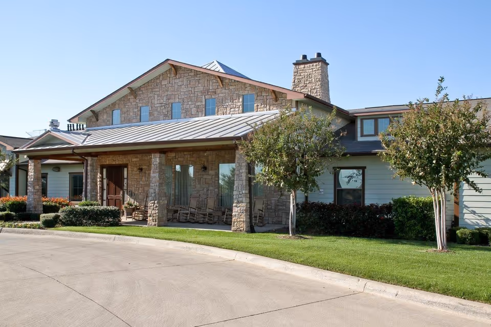 Front view of a stone-assisted living facility with porch seating and trees.