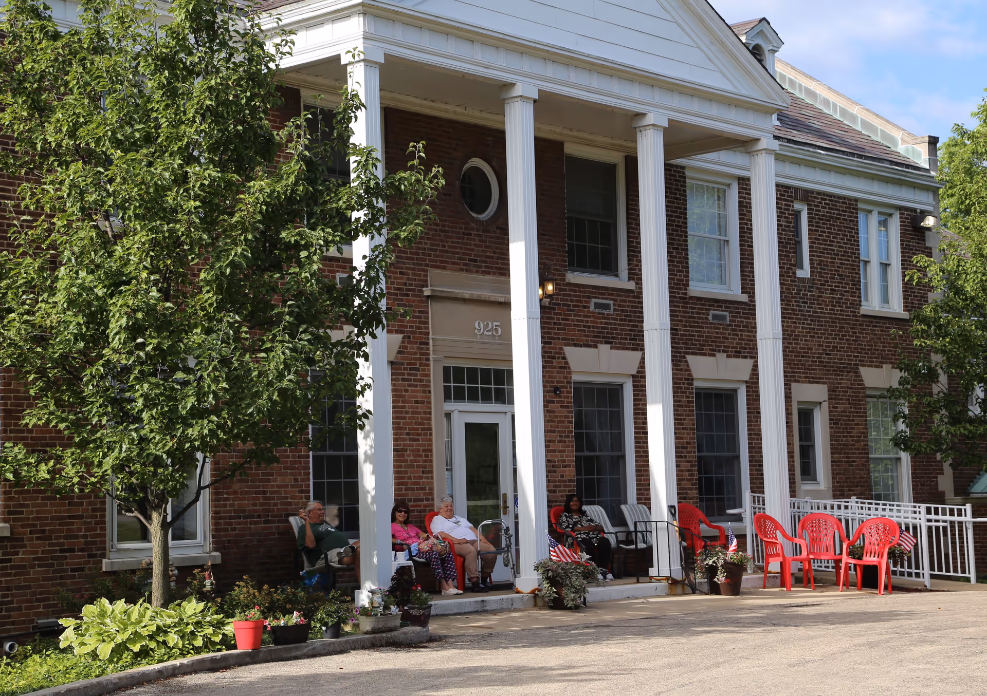 Front porch of a brick building with white columns and several people sitting on chairs enjoying the outdoor space. There are potted plants and small American flags decorating the area.