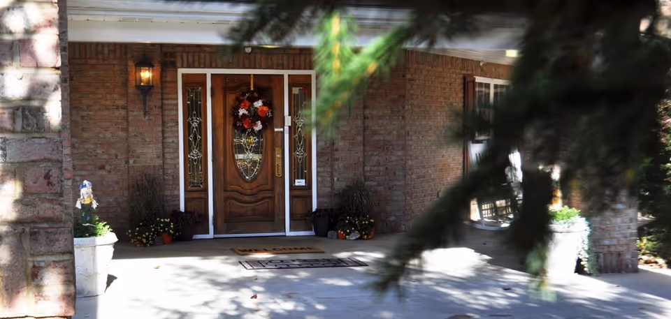 Front entrance of a brick building with a wooden door decorated with a fall-themed wreath. There are two welcome mats on the concrete porch, potted plants on either side of the door, and a wall-mounted lantern light fixture. Some tree branches partially obscure the view.