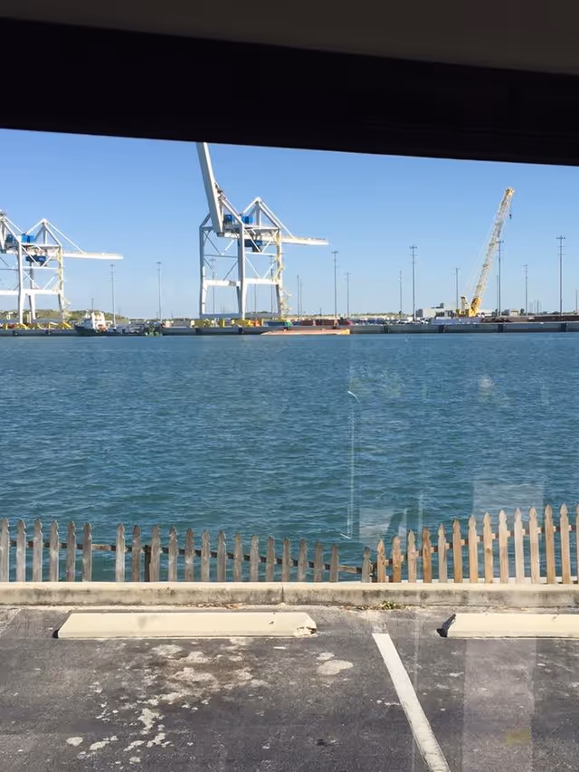 View of a waterfront industrial area with large cranes and a body of water, seen through a window above a parking lot with a wooden picket fence along the water's edge.
