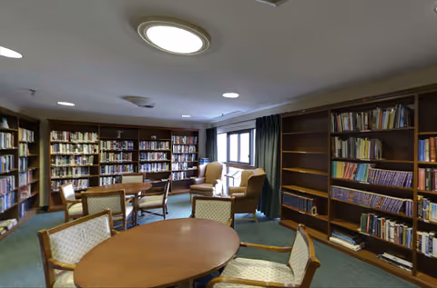 A cozy library room with wooden bookshelves filled with books along the walls, several round wooden tables surrounded by cushioned chairs, and two armchairs near a window with dark curtains. The room has a carpeted floor and ceiling lights.