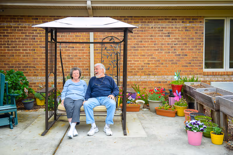An elderly couple sitting and smiling on a cushioned swing with a canopy in an outdoor patio area. They are surrounded by various colorful potted plants and flowers, with a brick wall and window in the background.