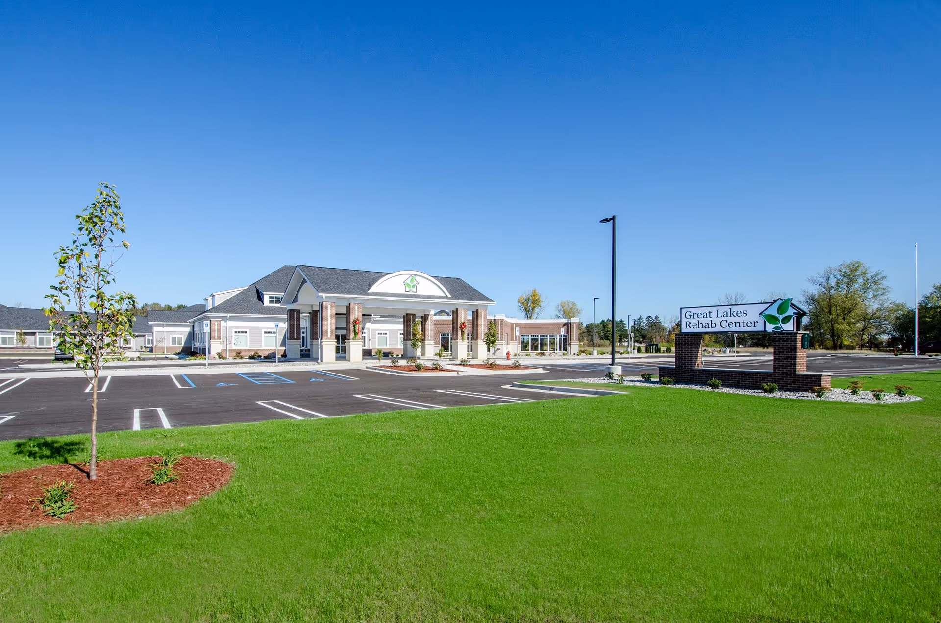 Exterior view of Great Lakes Rehab Center building with a large green lawn and a parking lot in front under a clear blue sky. A sign with the facility name is visible on the right side near the entrance.