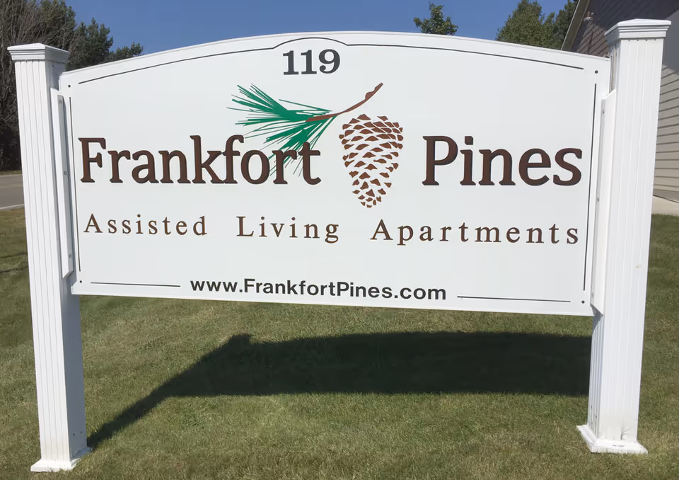 White outdoor sign reading "Frankfort Pines Assisted Living Apartments" with a pinecone logo and the website URL.
