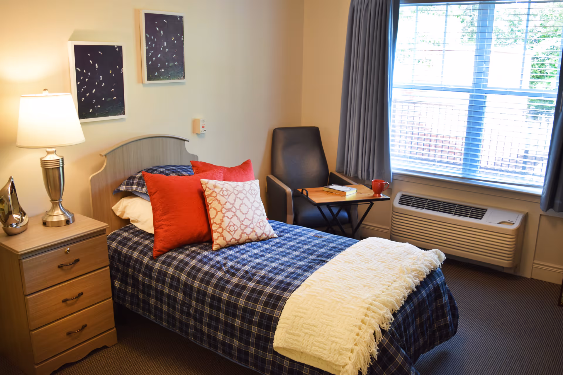 A tidy single bedroom with a plaid-covered bed, red accent pillows, a nightstand and lamp, an armchair, and a window with an air conditioning unit.