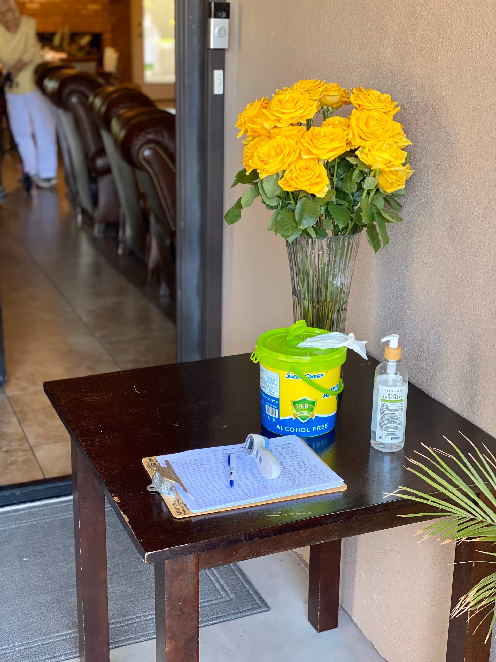 A dark entry table holds yellow roses, sanitizer, wipes, and a clipboard beside a doorway.