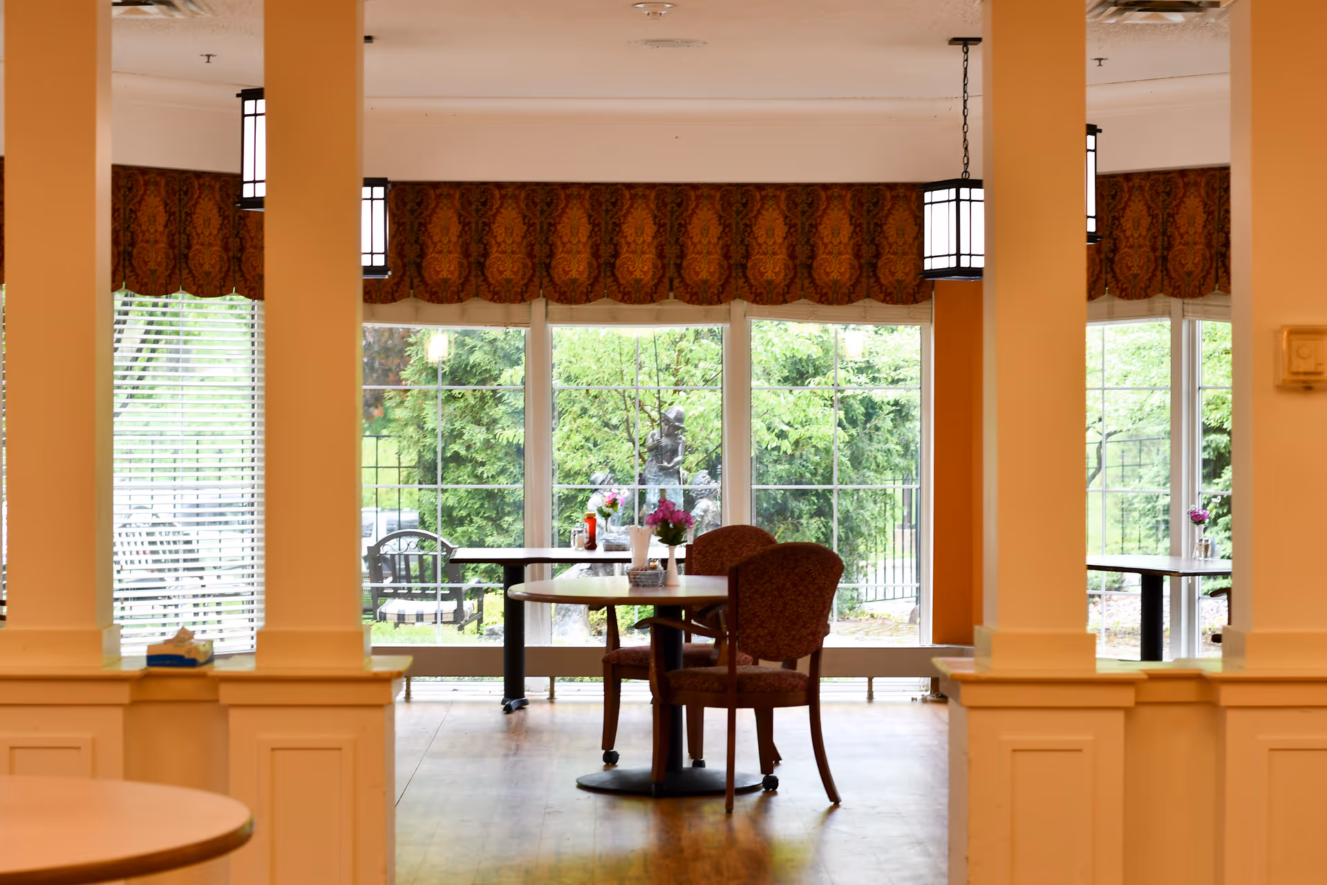 A dining area with round tables and upholstered chairs, large windows with a view of greenery outside, decorative valances above the windows, and hanging lantern-style light fixtures.