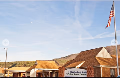 Exterior front of a brick senior care building with a sign reading 'Kissito Post Acute - The Brian Center' and an American flag against a blue sky.