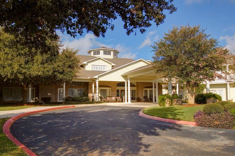 Senior living facility exterior with a covered entrance and landscaped driveway and trees.