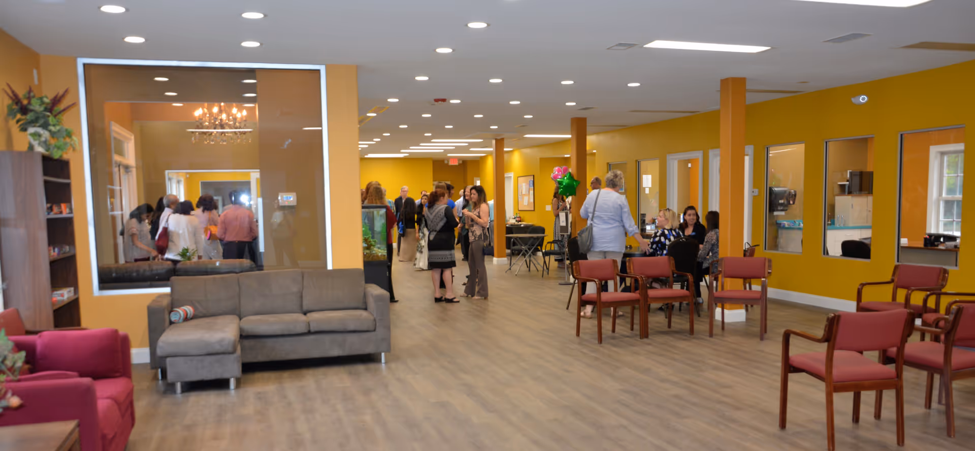 A spacious interior room with yellow walls and wood flooring, featuring a gray sectional sofa, red armchairs, and several wooden chairs arranged in small groups. People are standing and sitting, engaging in conversation. Large windows and glass panels separate this area from adjacent rooms, and there is a fish tank near the center.