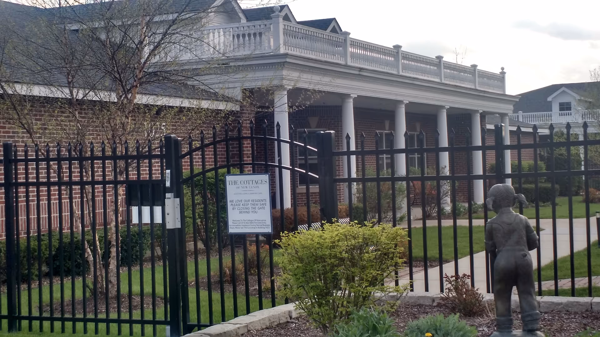 Brick senior living building with columned porch, black gate, and a statue.