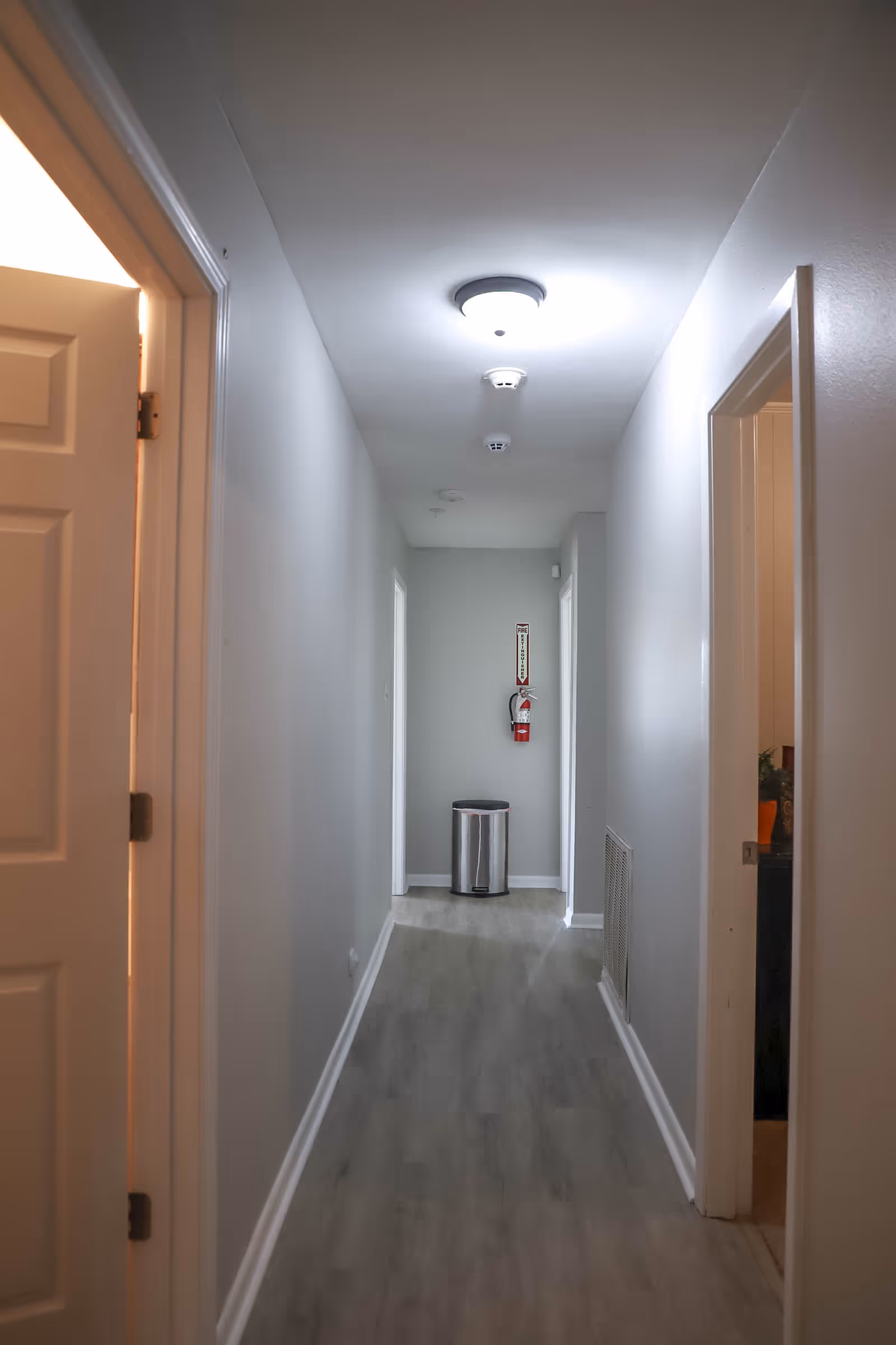 Narrow gray-painted hallway with ceiling lights, doors on both sides, and a trash can and fire extinguisher at the far end.