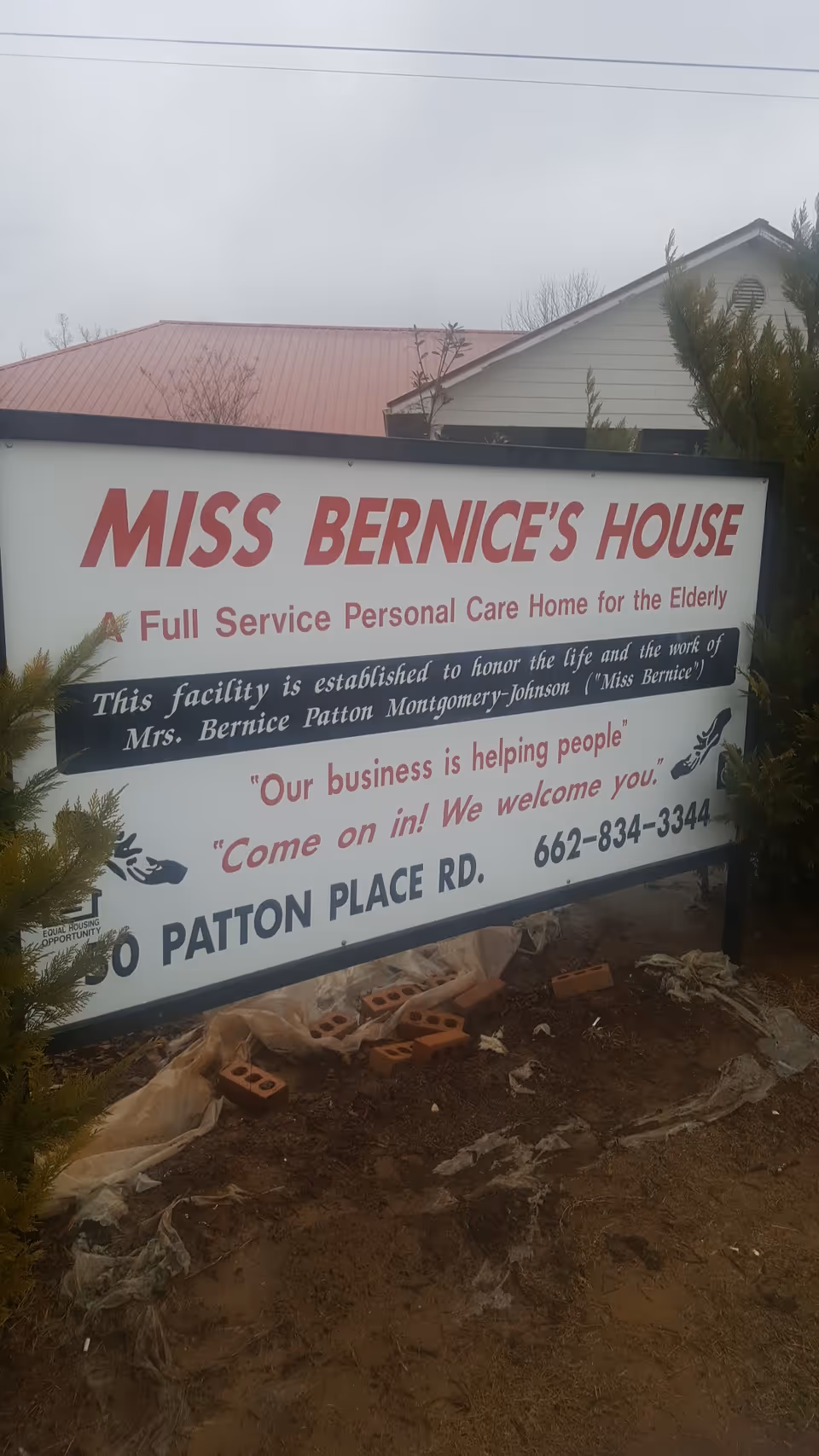Outdoor sign for Miss Bernice's House care home, with dirt and bricks nearby.