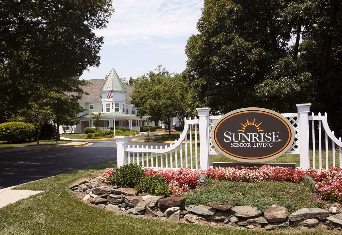 Entrance sign reading 'Sunrise Senior Living' in a landscaped flowerbed with the facility building and driveway in the background.