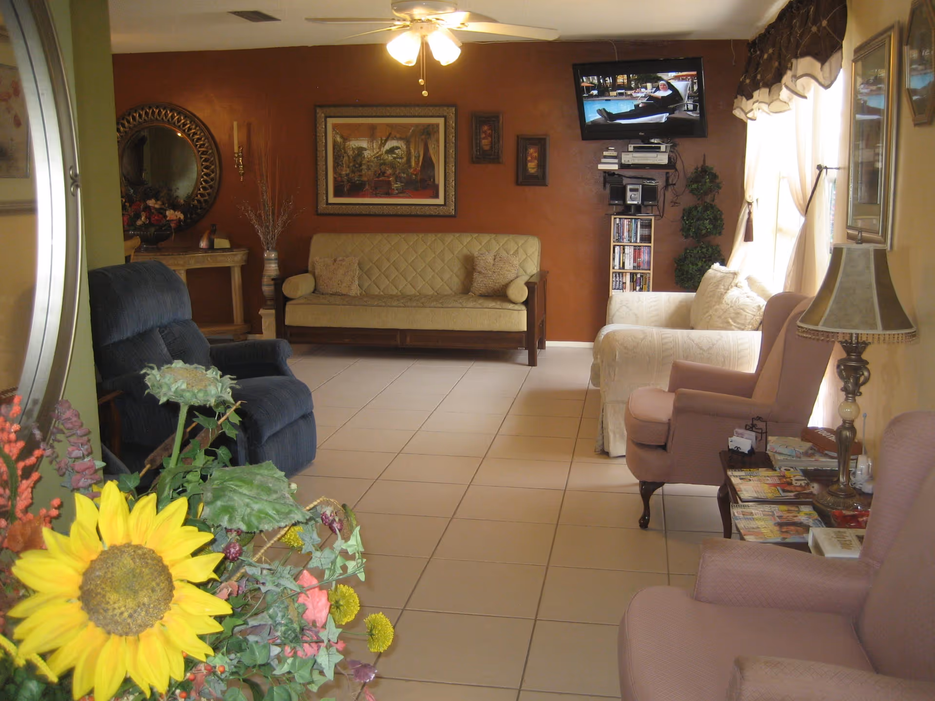 A cozy living room lounge with multiple armchairs and sofas, a wall-mounted TV, tiled floor, and a large sunflower arrangement in the foreground.
