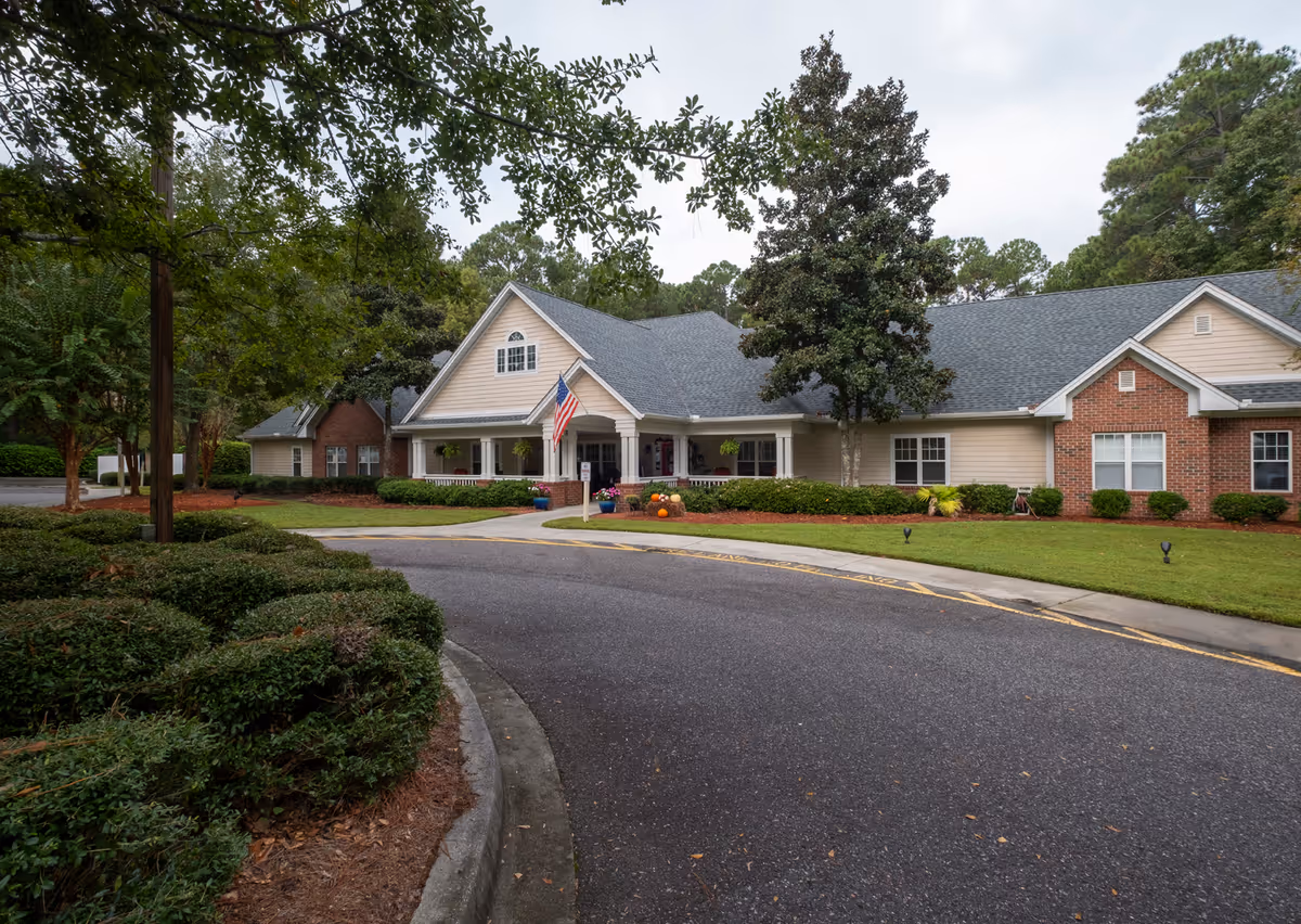 Exterior view of a single-story residential building with a gabled roof, an American flag near the entrance, surrounded by well-maintained landscaping including bushes, trees, and a curved driveway.