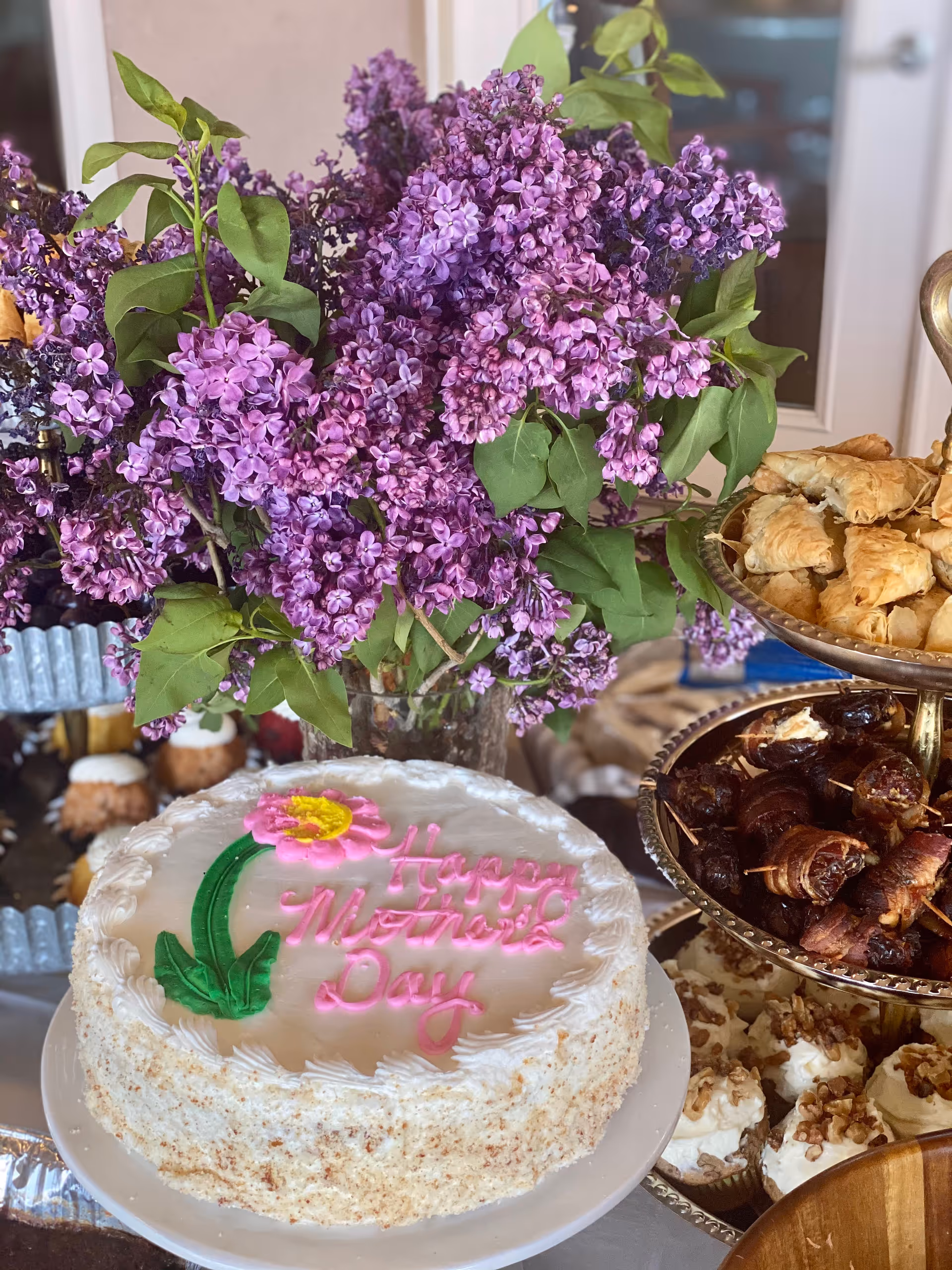 A cake with 'Happy Mother's Day' icing sits beside a purple lilac bouquet and pastries.