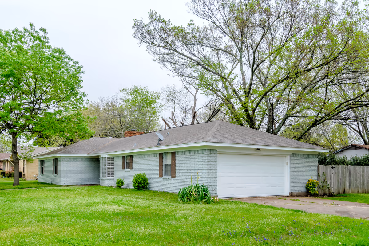 Single-story light-blue brick home with a two-car garage, green lawn, and trees.