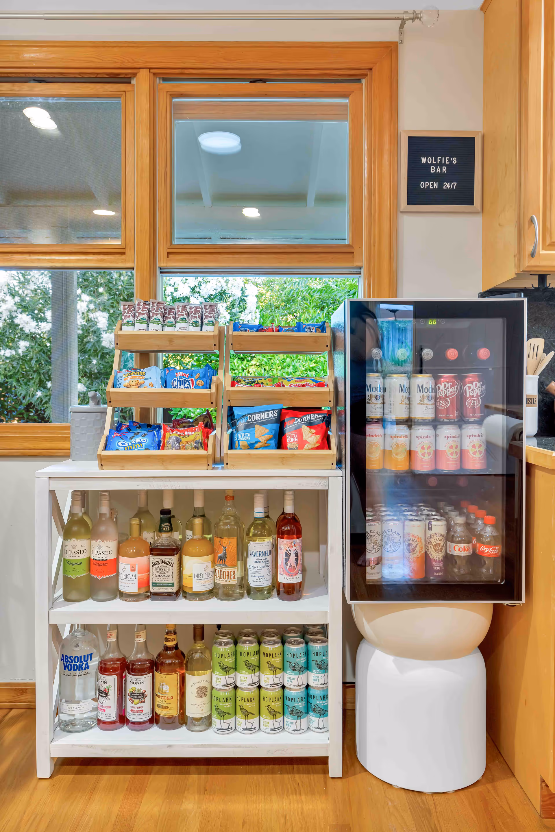 A small bar area named Wolfie's Bar with a white wooden shelf stocked with various alcoholic beverages and canned drinks. Above the shelf are two wooden racks filled with assorted snack packs. To the right is a glass-front mini fridge containing more canned and bottled drinks. The bar area is set against a wall with wooden window frames and a sign that reads 'WOLFIE'S BAR OPEN 24/7'.