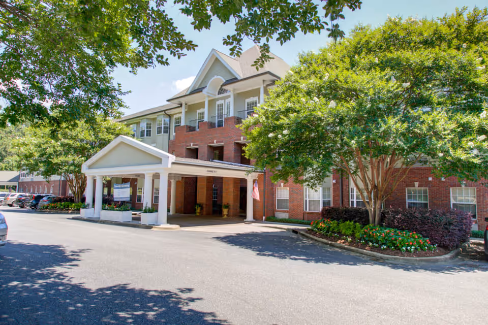 Front entrance of a brick senior living building with a covered portico, landscaping, and parked cars.
