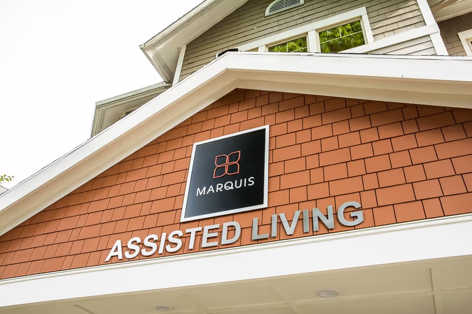 Close-up view of the exterior facade of Marquis Wilsonville Assisted Living facility, showing the building's name and logo on a brown shingle wall beneath a white roof overhang.