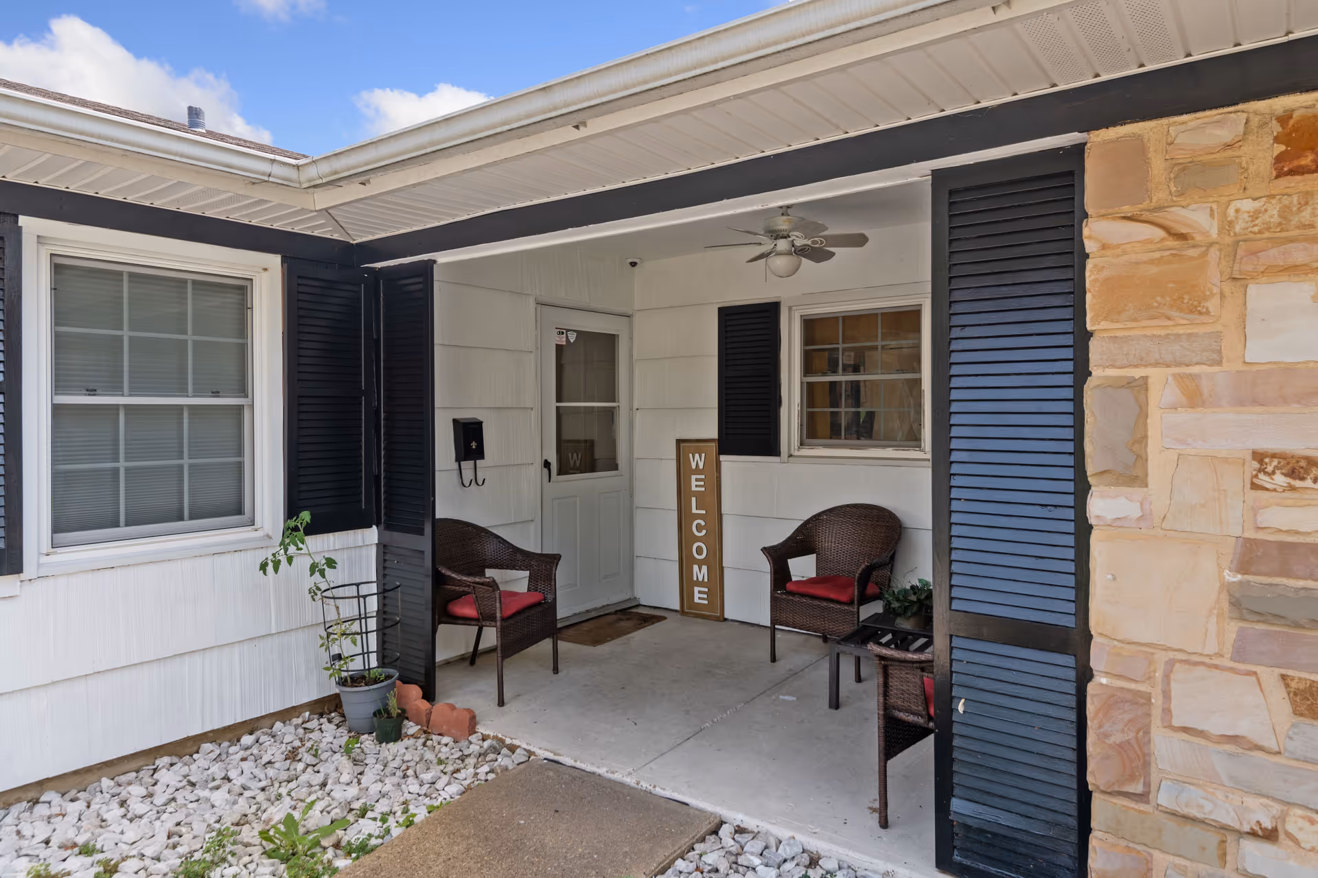 Covered front porch area of a residential building with two wicker chairs with red cushions, a small table, a welcome sign leaning against the wall, a ceiling fan, and a potted plant near the entrance door.