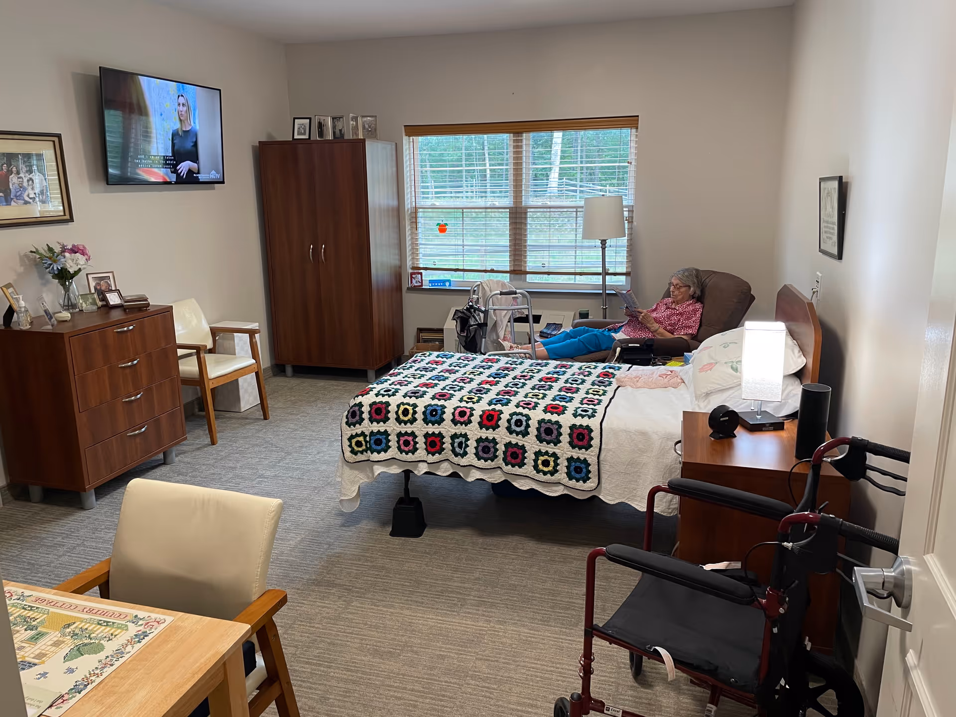 Senior resident relaxes in a furnished bedroom with bed, dresser, window, and TV.