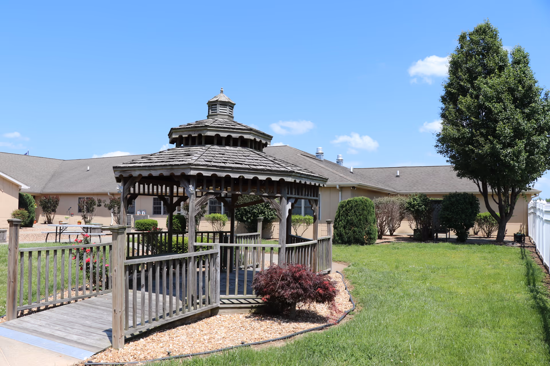 Outdoor view of Ash Grove Healthcare Facility & Rehabilitation showing a wooden gazebo with a shingled roof surrounded by a small garden area with bushes and a tree. The facility building is visible in the background under a clear blue sky.