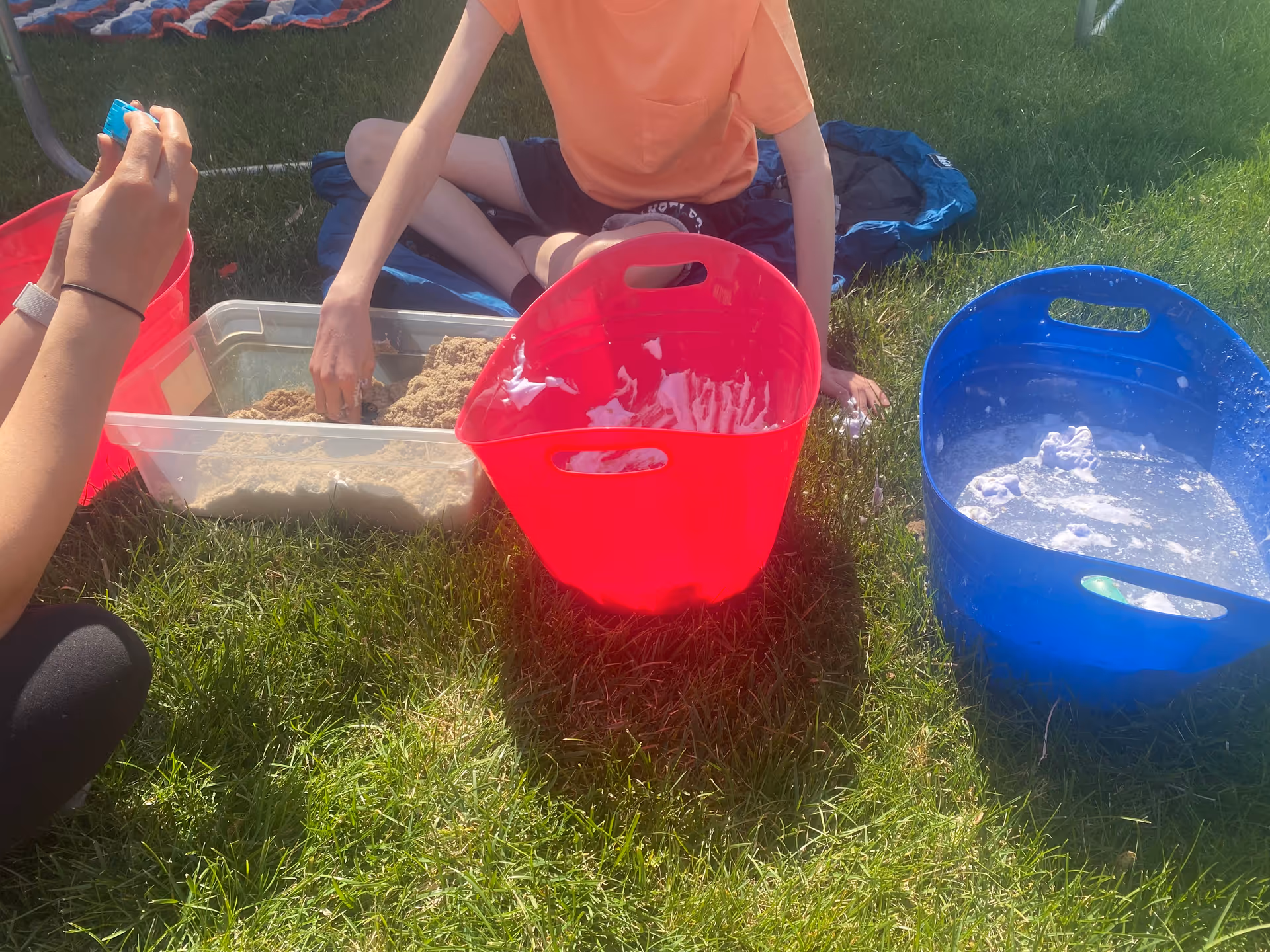 Two people sit on grass outdoors with sand, water, and colorful buckets.