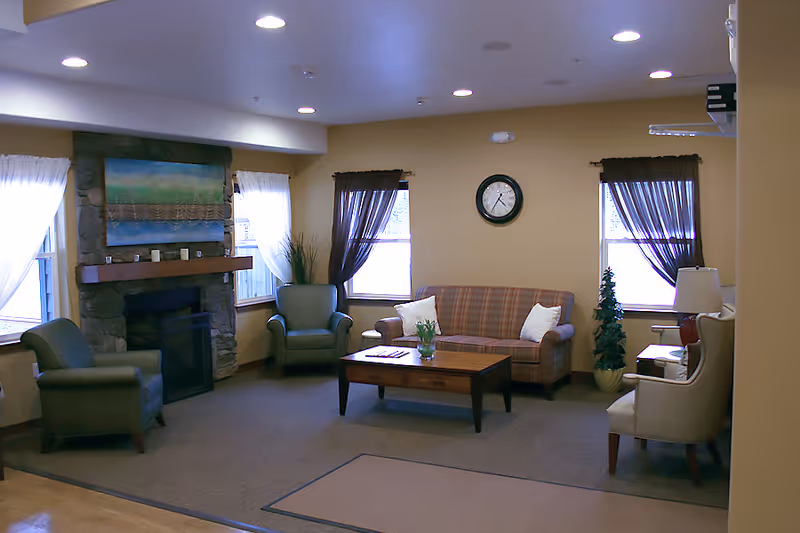A cozy living room with a stone fireplace, a plaid sofa, two green armchairs, a beige armchair, a wooden coffee table with a small plant, and windows with sheer and dark curtains.