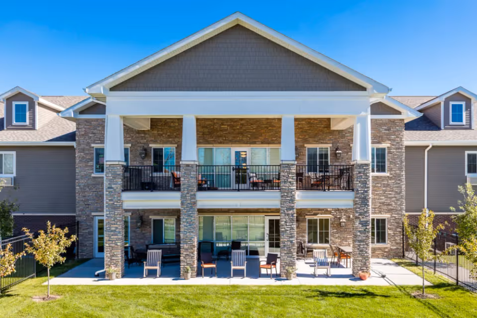 Front exterior of a two-story senior living building with stone facade, covered balconies and ground-level patio seating overlooking a lawn.
