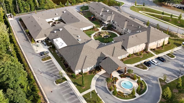 Aerial view of a senior living facility named Prema at Suwanee Creek, showing multiple connected buildings with a central courtyard, landscaped greenery, a circular driveway with a fountain, and surrounding parking areas.