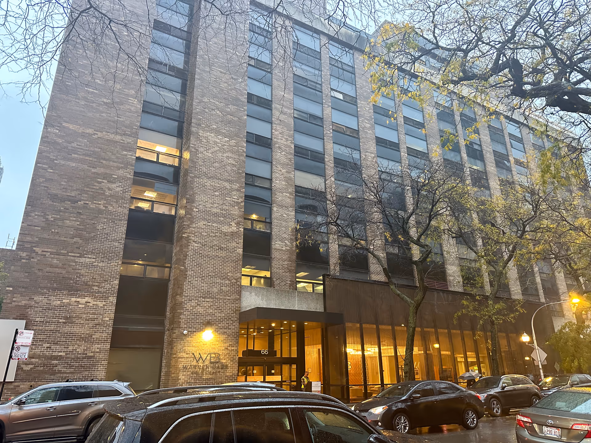 Front view of a multi-story brick residential building with a lit ground-floor entrance, trees along the sidewalk and parked cars on a wet street.