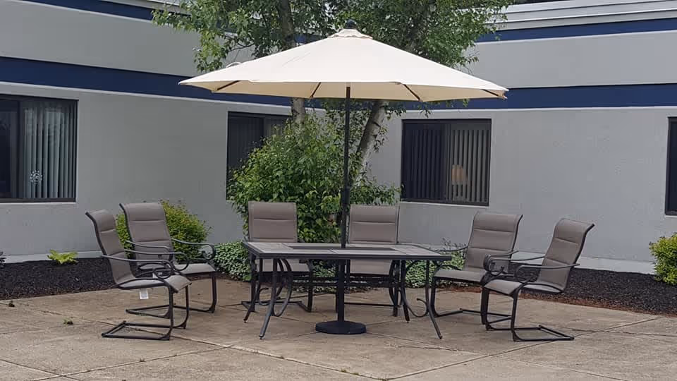 Outdoor patio area with a rectangular glass table, six cushioned chairs with metal frames, and a large beige umbrella providing shade. The patio is surrounded by a building with windows and some greenery including bushes and a tree.