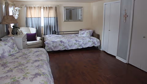A furnished senior living facility bedroom with two floral beds and a seating area.