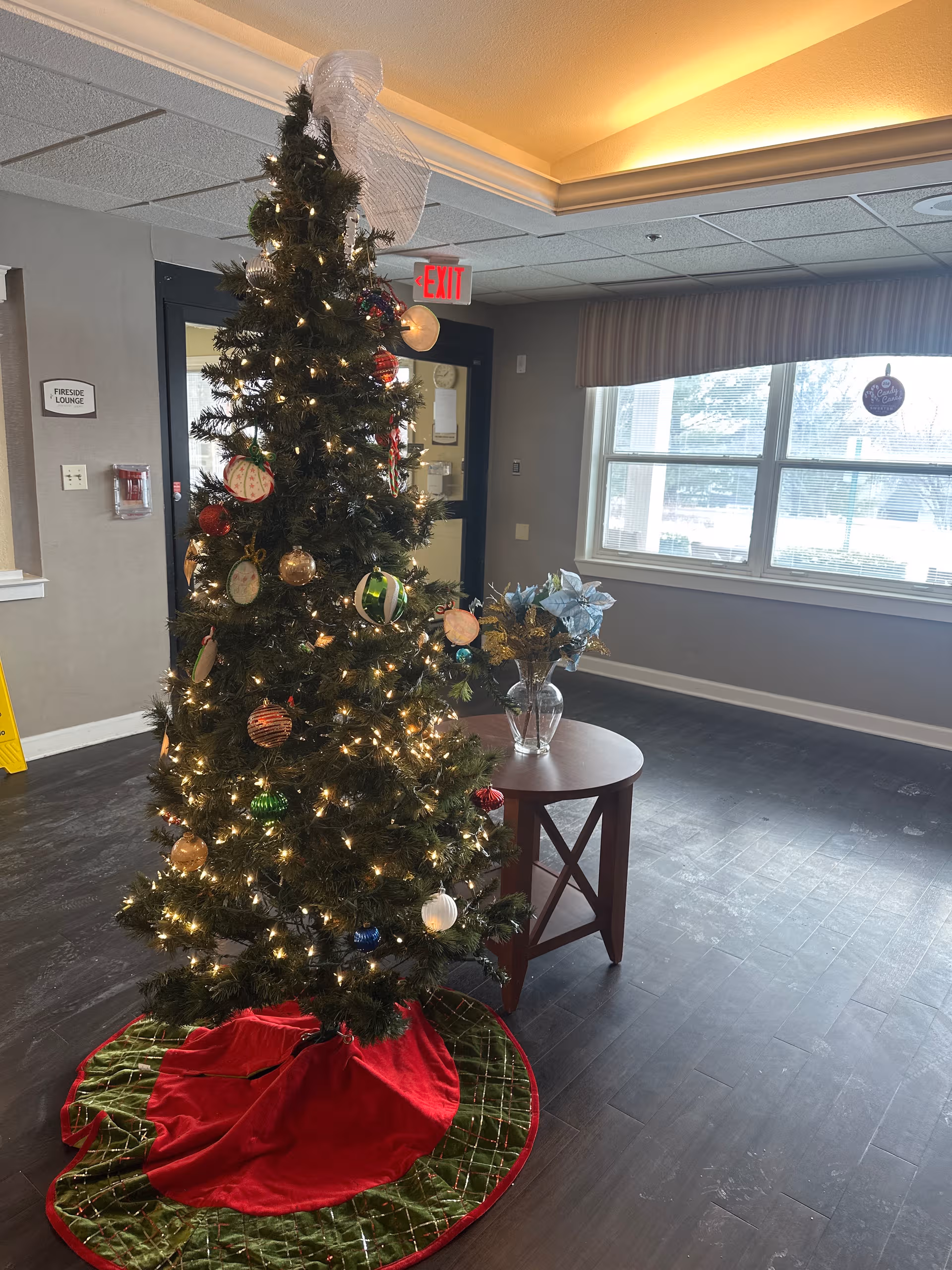 Holiday decorated Christmas tree in a facility lobby with a small table and windows.