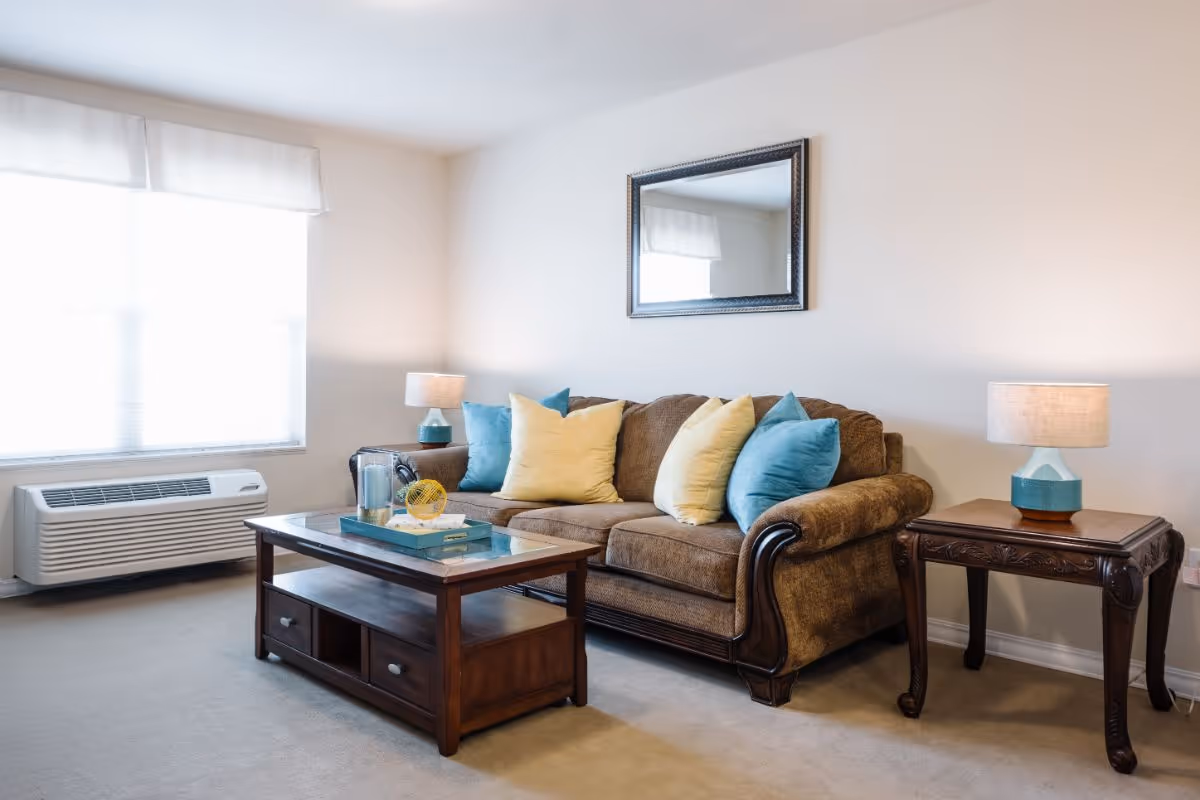 A cozy living room with a brown upholstered sofa adorned with yellow and blue throw pillows. In front of the sofa is a wooden coffee table with a glass top, holding a decorative tray with a pitcher, glass, and a small yellow decorative object. On either side of the sofa are wooden side tables with matching blue and white lamps. A large window with white curtains lets in natural light, and a framed mirror hangs on the wall above the sofa.