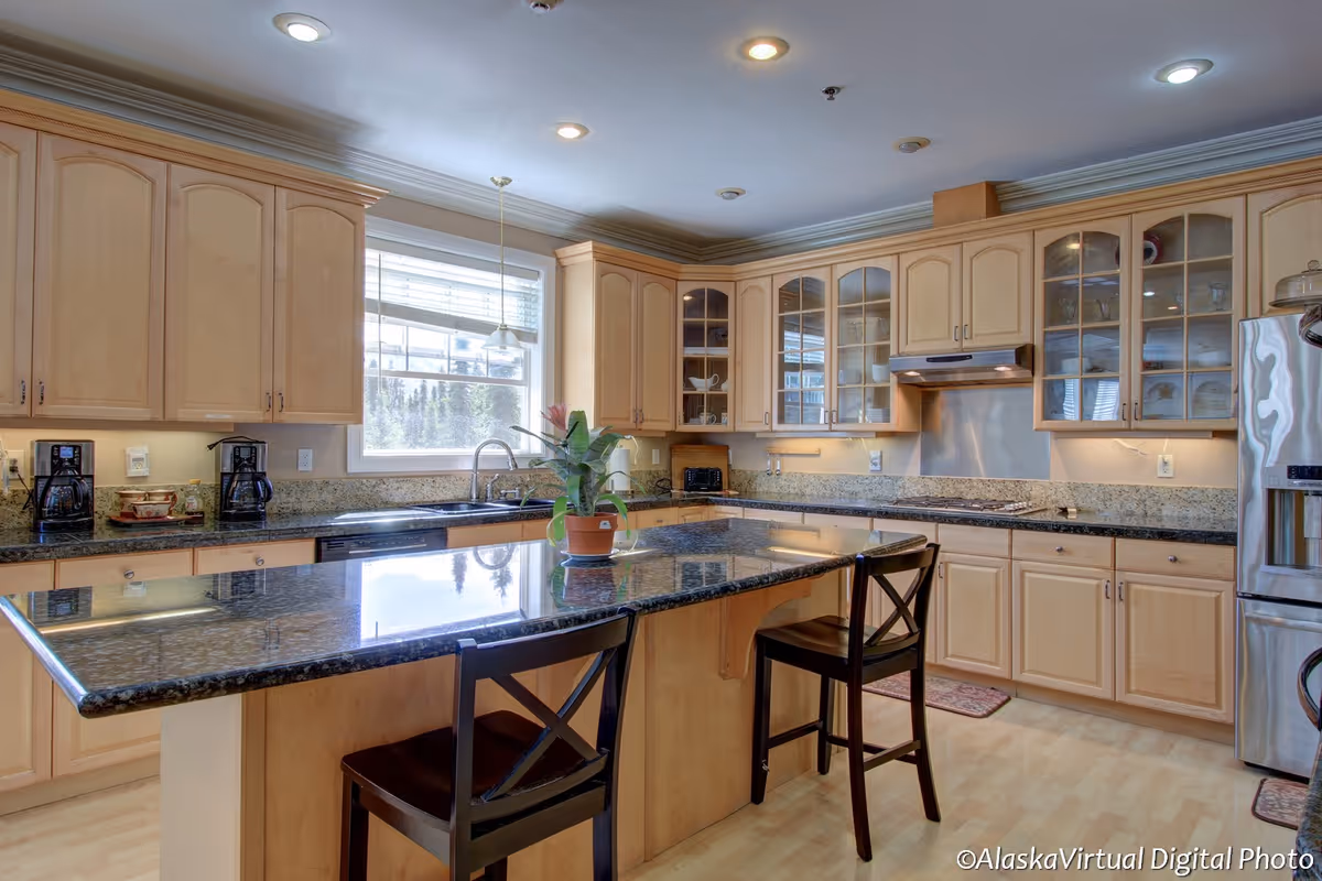 A spacious kitchen with light wood cabinets, granite countertops, and a large island with two dark wooden chairs. The kitchen features a stainless steel refrigerator, a stove with a hood, a dishwasher, a double sink under a window, and various small appliances like coffee makers and a toaster. There is a potted plant on the island and recessed lighting in the ceiling.