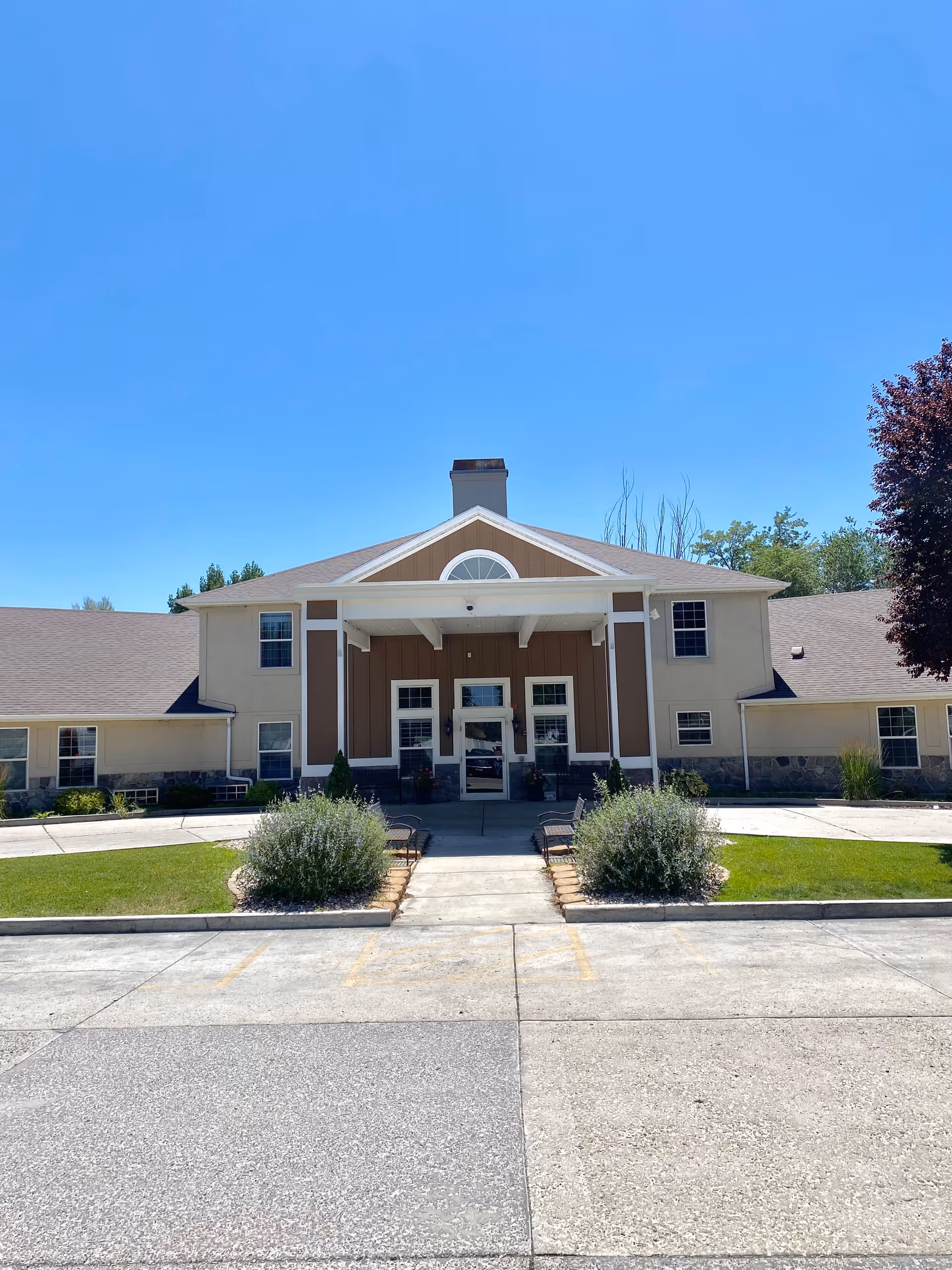 Front exterior view of a senior living facility building with a covered entrance, multiple windows, and a well-maintained lawn with shrubs and benches on either side of the walkway leading to the entrance. The sky is clear and blue.