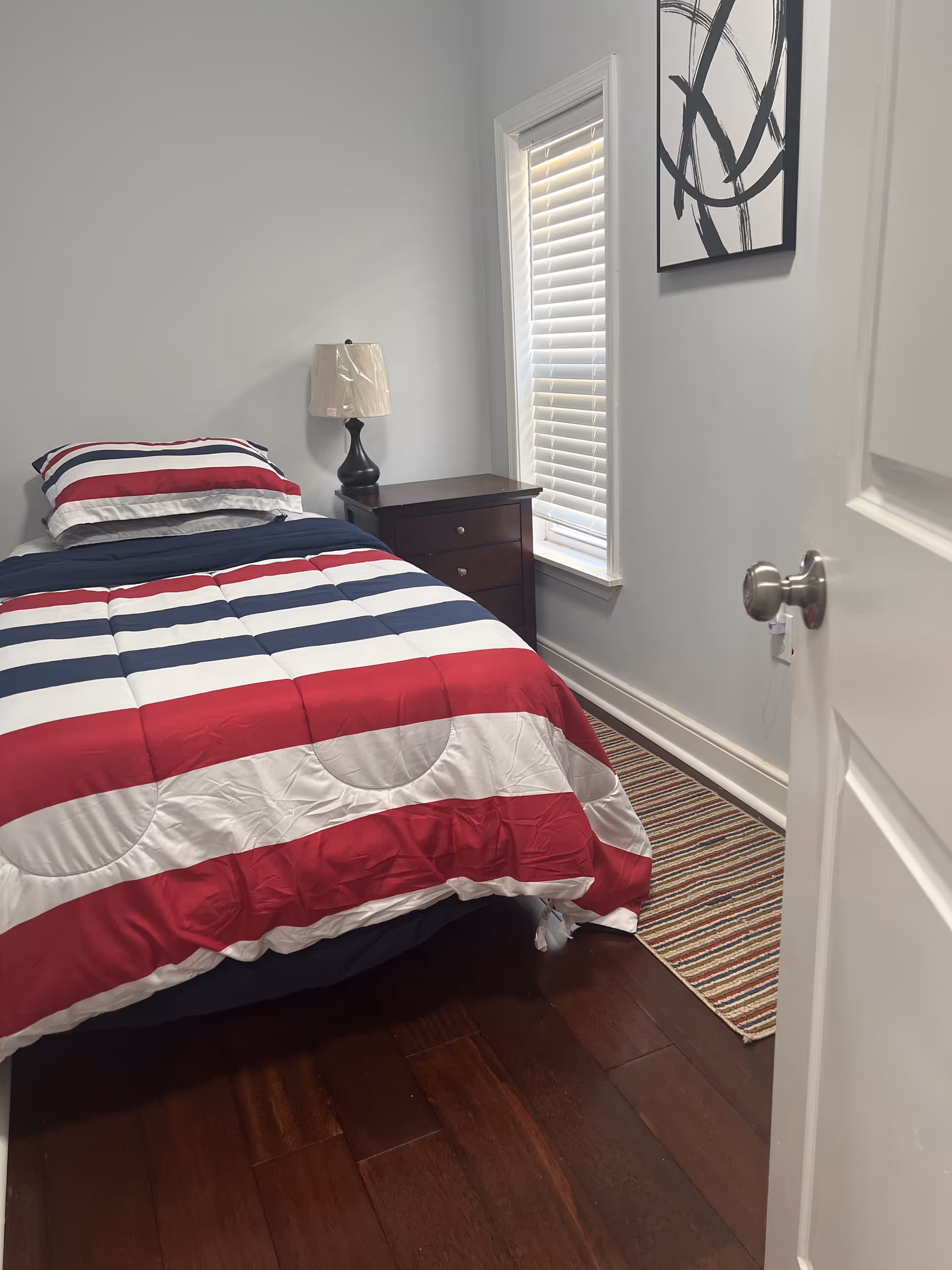 A small bedroom with a striped red, white, and blue bedspread, nightstand, lamp, and window blinds.
