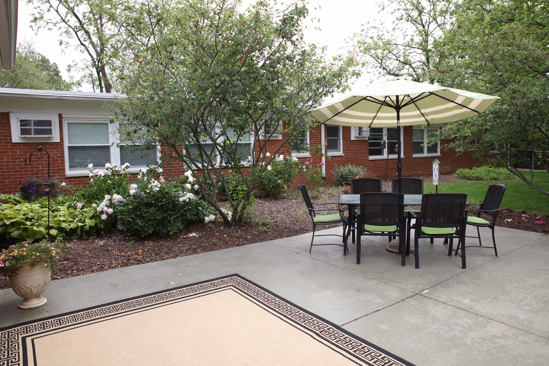 Outdoor patio courtyard with a table, umbrella and chairs in front of a single-story brick building surrounded by trees and flowerbeds.